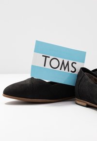 Černé perforované baleríny s špičatou špičkou. Hnědá gumová podrážka. Štítek s nápisem "TOMS" tučnými černými písmeny na světle modrém pozadí.