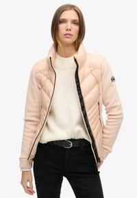 Lys pink quilted jakke med høj krave og sort lynlås; har et logo patch på skulderen og sorte detaljer. Bæres over en hvid sweater.