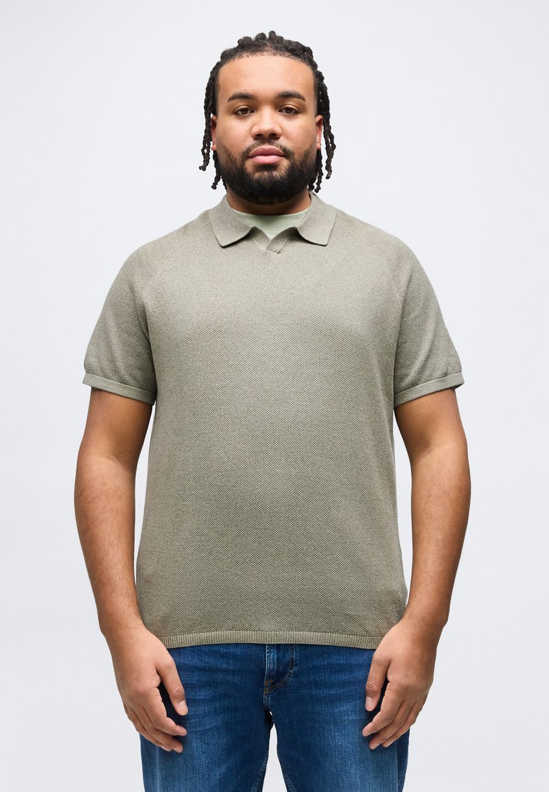 Άνδρας με πλεξούδες στα μαλλιά και γένια, φοράει γκρι πλεκτό πόλο και μπλε τζιν, στέκεται μπροστά σε απλό λευκό φόντο.