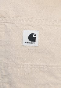 Beige canvas tyg med en fyrkantig vit etikett som har en svart logotyp. Synliga sömmar och texturdetaljer är närvarande.