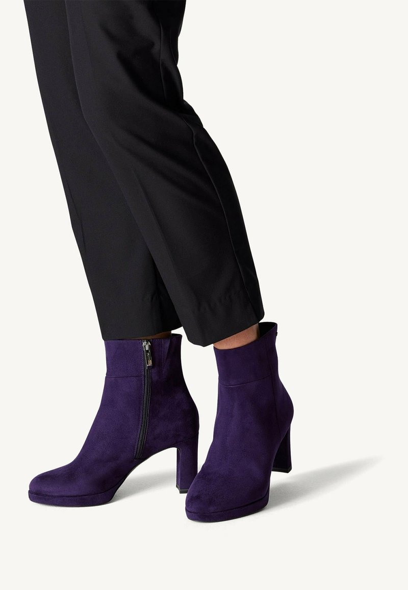 Personne portant un pantalon noir et des bottines violettes en daim avec des talons blocs et des fermetures éclair latérales visibles, debout sur une surface claire.