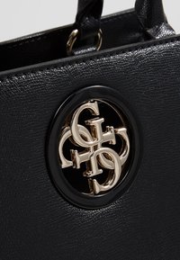 Sac à main noir texturé avec un emblème rond en or présentant les lettres entrelacées G et H sur le devant.