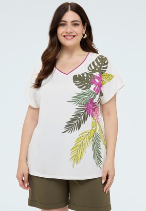 Mujer sonriente con cabello oscuro lleva una blusa blanca con estampado colorido de hojas tropicales y pantalones cortos verde oliva sobre un fondo claro y liso.