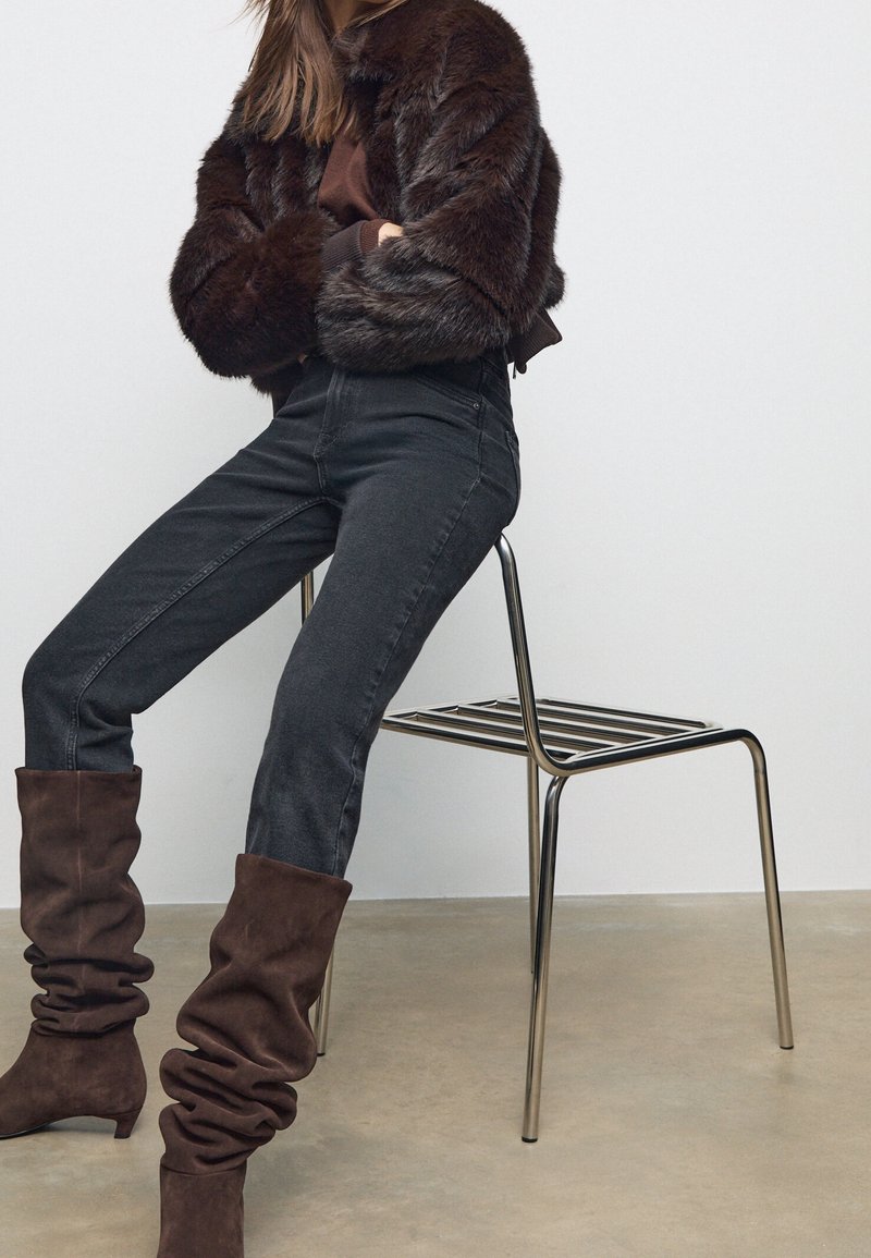 Femme portant une veste en fourrure foncée, des bottes marron décontractées et un jean noir, assise sur une chaise en métal sur un fond uni.