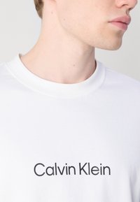 Balta medvilninė marškinėlių su apvali apykakle ir juodu „Calvin Klein“ logotipu, atspausdintu ant krūtinės. Lygus paviršius, paprastas dizainas.