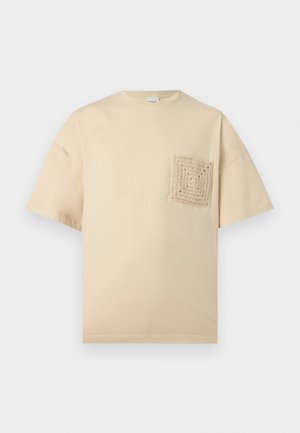 Man with curly hair wearing black sunglasses, beige oversized t-shirt with textured pocket, loose taupe trousers, and black dress shoes.