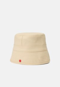 Beige kbelíková čepice vyrobená z umělého materiálu, se hladkým povrchem, prošívanými detaily a malou červenou emblémem na boku.