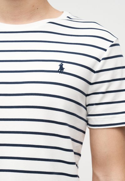 Λευκό μπλουζάκι με ναυτικά οριζόντια ρίγες που φοριέται από άτομο, με ένα μικρό κεντητό λογότυπο στο αριστερό στήθος.