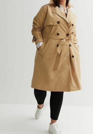 Personne portant un trench-coat beige avec ceinture, pantalon noir, baskets blanches, les mains dans les poches du manteau sur un fond uni.