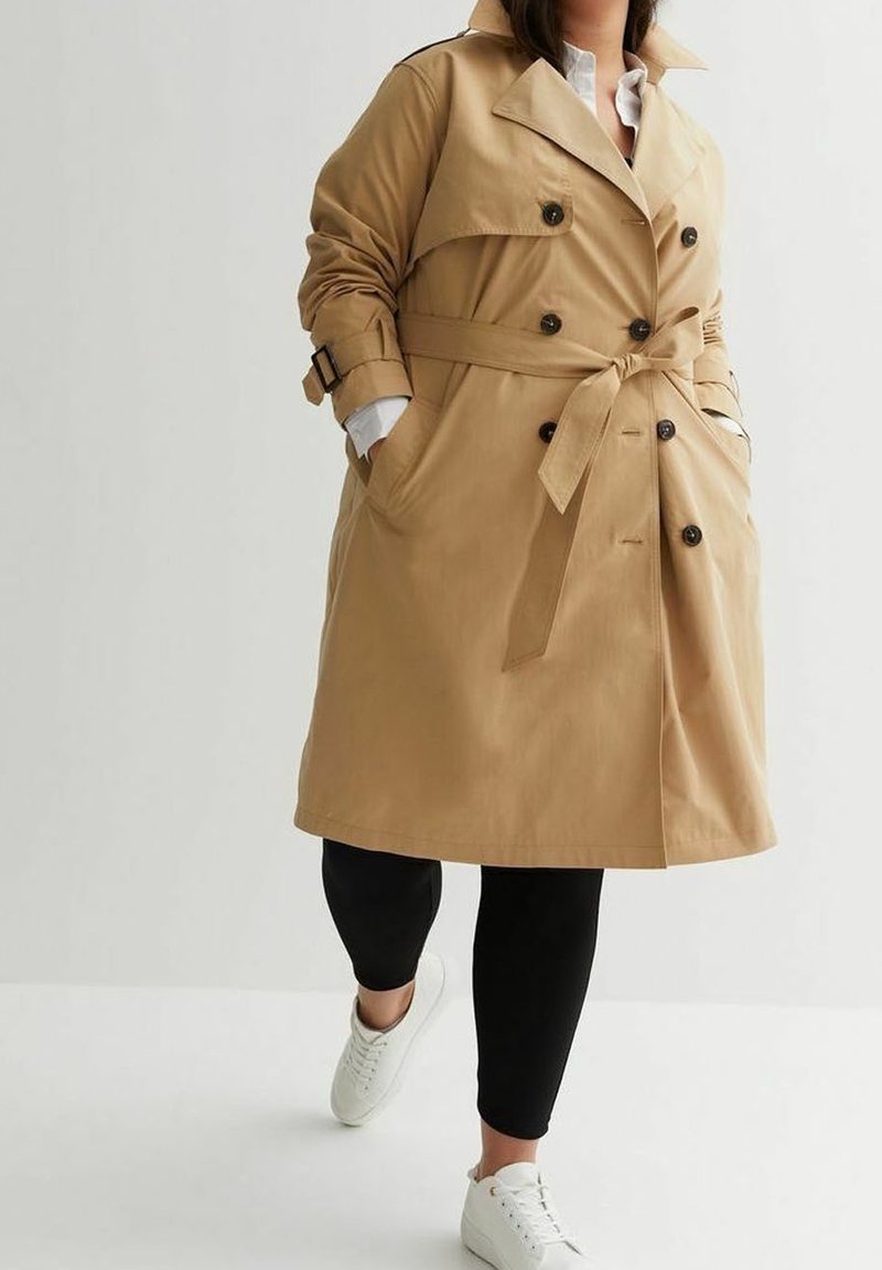 Personne portant un trench-coat beige avec ceinture, pantalon noir, baskets blanches, les mains dans les poches du manteau sur un fond uni.