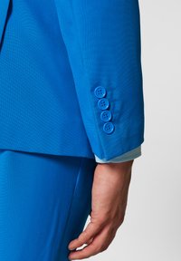 Erksinine ülikonna jakk, millel on kohandatud sobivus, sile tekstuur ja neli sinist nööpi varrukal koos nähtavate õmbluse detailidega.