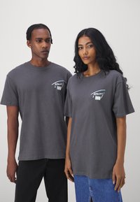 Camisetas de algodón gris con mangas cortas. El bolsillo delantero cuenta con un diseño gráfico colorido. Modelo a la izquierda con jeans negros, a la derecha con falda de mezclilla.