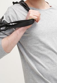 Grå långärmad tröja med rund halsringning, tillverkad i mjukt material, bärs medan man håller i ett svart rem av en väska med strukturerad yta.