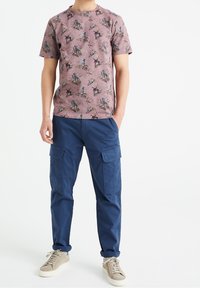Mężczyzna w fioletowej koszulce w kwiatowy wzór, niebieskich spodniach cargo z kieszeniami i beżowych trampkach, stojący z jedną ręką w kieszeni.