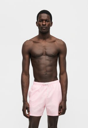 Μυώδης άνδρας που στέκεται με το πρόσωπο προς τα εμπρός, φοράει ανοιχτό ροζ μαγιό με κορδόνι, σε απλό γκρι φόντο.