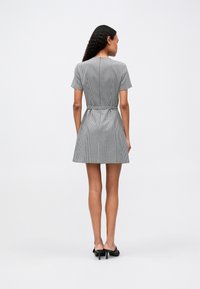 Γυναίκα με μακριά σγουρά μαλλιά, φορούσε κο�ντό  manches φόρεμα σε λευκό και μαύρο καρό σχέδιο και μαύρα ψηλοτάκουνα μules, στέκεται μπροστά σε λευκό φόντο.