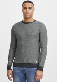 Gray long-sleeve sweater made of textured knit fabric with a black ribbed neckline and hem, featuring a small logo patch at the bottom.