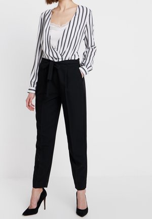 Femme portant un chemisier blanc à rayures verticales noires, un pantalon noir taille haute avec une ceinture en tissu, et des chaussures à talons hauts noires.