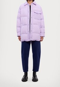 Chaqueta acolchada de un ligero color púrpura con botones a presión, bolsillo en el pecho y un corte holgado, llevada sobre una camiseta negra, combinada con pantalones anchos azul marino y botas.