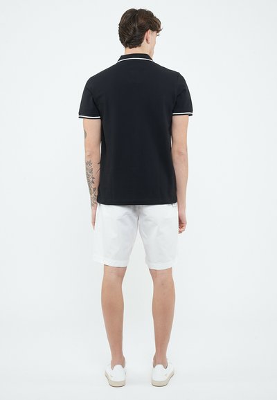 Man die met zijn rug naar de camera staat, gekleed in een zwart poloshirt met korte mouwen en witte bies, witte knielange korte broek, witte sneakers en een zichtbare armtattoo.