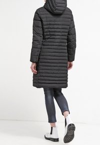 Long black puffer coat with hoodie, quilted pattern, and side seams; paired with blue jeans and white ankle boots with black detailing.