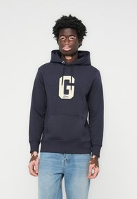 Marineblå hættetrøje med et hvidt "G" logo, snøre i hætten, forlomme, ribbede manchetter og kant. Blødt stof med et afslappet design.