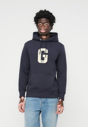 Marineblå hettegenser med en hvit "G"-logo, snorhette, frontlomme, ribbestrikkede mansjetter og hem. Myk stoffkvalitet med et uformelt design.