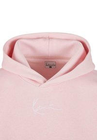 Lys rosa hettegenser med stor hette, myk stofftekstur, og en hvit brodert logo på forsiden. Innvendige sømmer er pent utført.