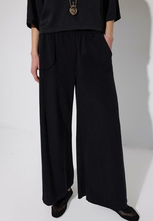 Persoon die losse zwarte broek met een elastische tailleband draagt, zwarte schoenen en een zwart topje met een hangerketting, handen in de zakken.