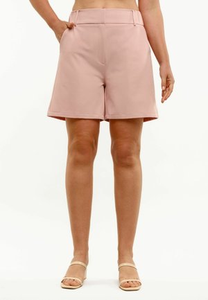 Kvinde iført lyserøde skræddersyede shorts med høj talje og lommer samt beige åbne hæle, stående mod en hvid baggrund.