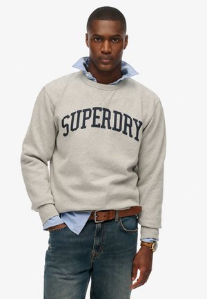 Grijze fleece sweatshirt met "SUPERDRY" in donkerblauwe letters, gedragen over een lichtblauw overhemd met kraag, gecombineerd met blauwe jeans en een leren bruine riem.