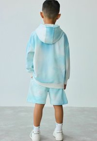 Sudadera azul clara con acentos blancos, combinada con pantalones cortos a juego. Textura suave, corte holgado y un sutil patrón parecido a nubes. Zapatillas blancas.