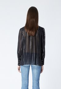 Blusa negra transparente con rayas verticales blancas, mangas largas, puños abotonados y dobladillo recto, combinada con pantalones vaqueros de color azul claro.