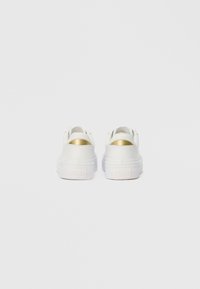 Een paar witte sneakers met gouden hielaccenten, gezien vanaf de achterkant, op een effen lichtgrijze achtergrond.