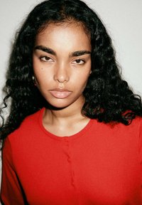 Jeune femme aux longs cheveux noirs bouclés portant un haut rouge boutonné, regardant directement la caméra sur un fond uni.