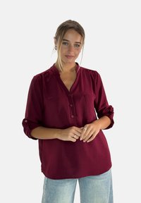 Camicia bordeaux con maniche a tre quarti, taschino frontale e finitura texturizzata, abbinata a jeans azzurri a vita alta.