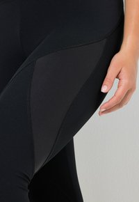 Czarne legginsy mają prążkowaną fakturę, bezszwowy design oraz lekko błyszczący panel boczny. Dłoń spoczywa na udzie dla porównania rozmiaru.