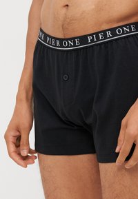Boxers en coton noir avec un devant à boutons et une ceinture comportant "PIER ONE" en texte blanc sur une rayure grise. Texture douce avec une coupe ample.