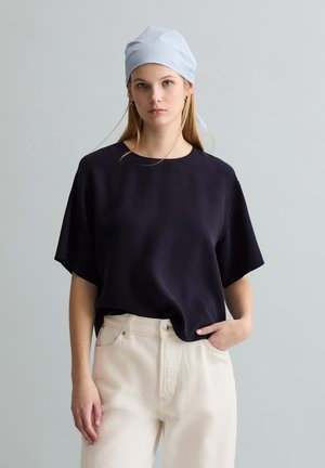 Junge Frau mit langen blonden Haaren, die ein gestreiftes Kopftuch, ein schwarzes Kurzarmshirt und cremefarbene, hoch taillierte Hosen trägt, stehend vor einem grauen Hintergrund.