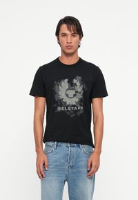 Črna bombažna majica s sivim grafičnim logotipom, okroglim izrezom, kratkimi rokavi in običajnim krojem. Obuta je z svetlo modrimi jeans hlačami.