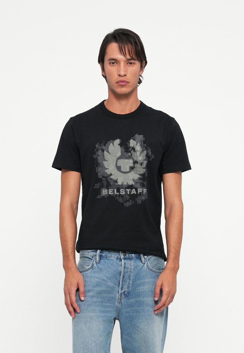 Črna bombažna majica s sivim grafičnim logotipom, okroglim izrezom, kratkimi rokavi in običajnim krojem. Obuta je z svetlo modrimi jeans hlačami.