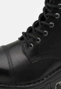 Botte en cuir noir avec bout rond, cap renforcé, œillets en métal et lacets noirs. Présente une surface texturée et un détail de couture contrasté.
