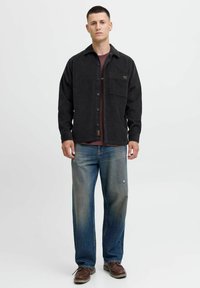 Camisa de pana negra con botones, bolsillo frontal, usada sobre una camiseta de color burdeos. Jeans azul desgastados, zapatos marrones con suelas claras.