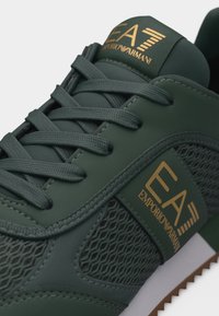 Σκούρο πράσινο αθλητικό παπούτσι EA7 Emporio Armani με υφή από δίχτυ και δέρμα, χρυσό λογότυπο στο πλάι και στη γλώσσα, και κορδόνια σε ταιριαστό πράσινο χρώμα.