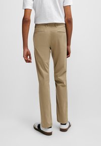 Chino en khaki avec un devant plat, deux poches arrière et une fermeture éclair. Porté avec une chemise blanche à manches courtes et des baskets blanches avec des accents noirs.