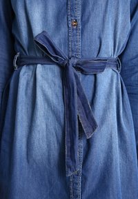 Denimová košilová šaty s zapínáním na knoflíky, dlouhými rukávy a tmavším denimovým páskem uvázaným v pase. Má měkkou texturu a rovnoměrnou modrou barvu.