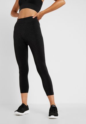Femme portant des leggings noirs courts et des baskets noires, debout sur un fond clair uni.