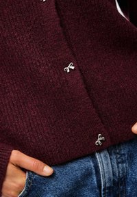 Burgundijas krāsas adīts kardigāns ar maziem sudraba lentīšu akcentiem, teksturēta auduma, un ribotiem piedurknēm, saskaņots ar ziliem džinsu biksēm.
