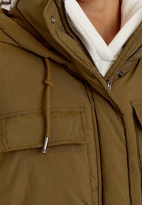 Grüne Kapuzenjacke mit strukturiertem Finish, ausgestattet mit einem Reißverschluss, zwei Brusttaschen und verstellbarem Kordelzug. Futter aus weißem Innenmaterial.