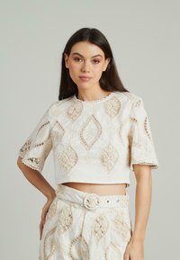 Witte cropped top met ingewikkelde kantdetails, voorzien van diamanten patronen en gegolfde randen. Korte mouwen complementeren het textuurontwerp.